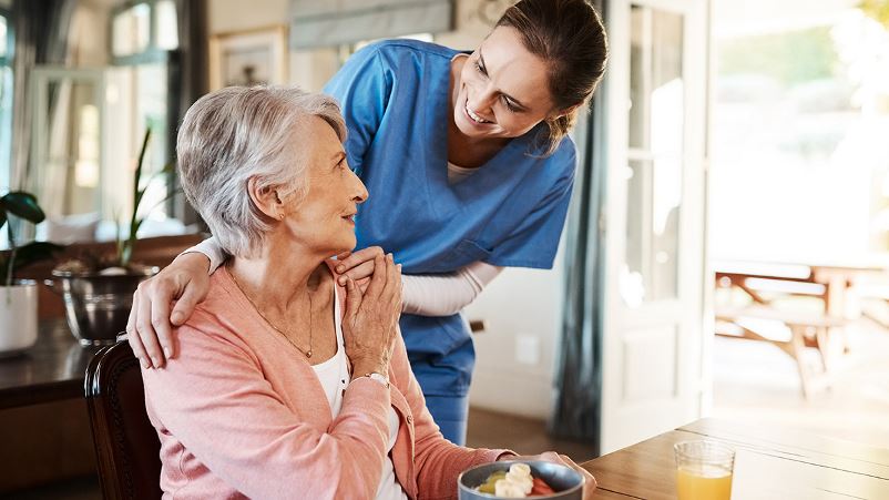 Older adult woman eating a healthy breakfast and being visited by a health care professional.